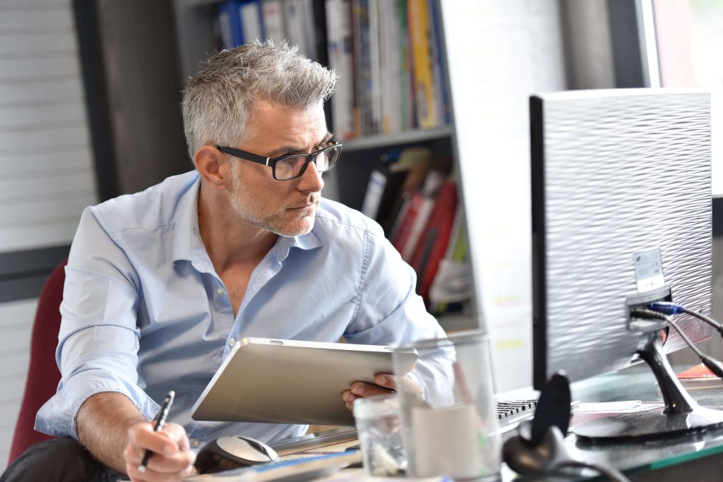 Ein Mann mit grauen Haaren und Brille sitzt an einem Schreibtisch vor einem Computer, hält ein Tablet in der einen und einen Stift in der anderen Hand und schaut auf den Bildschirm.